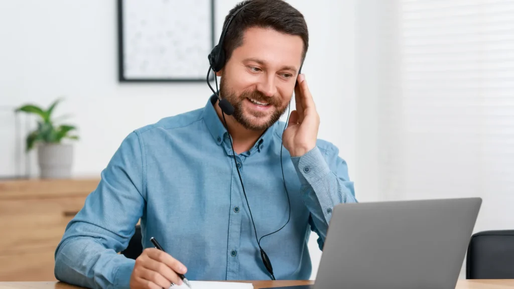 Homem utilizando fone de ouvido com microfone e notebook durante uma conversa remota, ilustrando o uso de tecnologias de tradução simultânea para comunicação entre diferentes idiomas.
